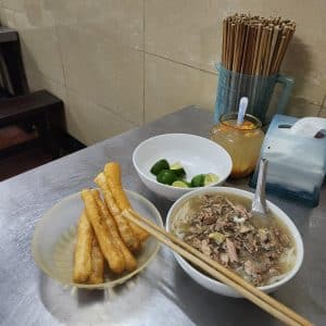 A bowl of Pho with no onion leaves and bowls of lemon chilli vinegar and bread sticks.